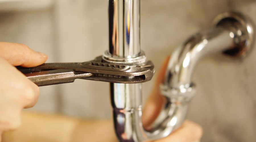 Person using a wrench to tighten a chrome plumbing pipe, emphasizing the importance of regular plumbing maintenance to prevent issues and ensure system efficiency.