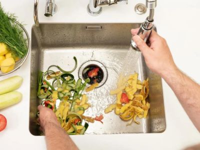 Hands rinsing vegetable scraps in a kitchen sink with a garbage disposal, emphasizing proper food waste disposal practices to prevent clogs.